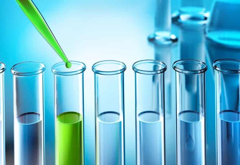 A green liquid is being dropped from a pipette into one of several clear test tubes, with the surrounding lab equipment blurred in the blue-toned background.