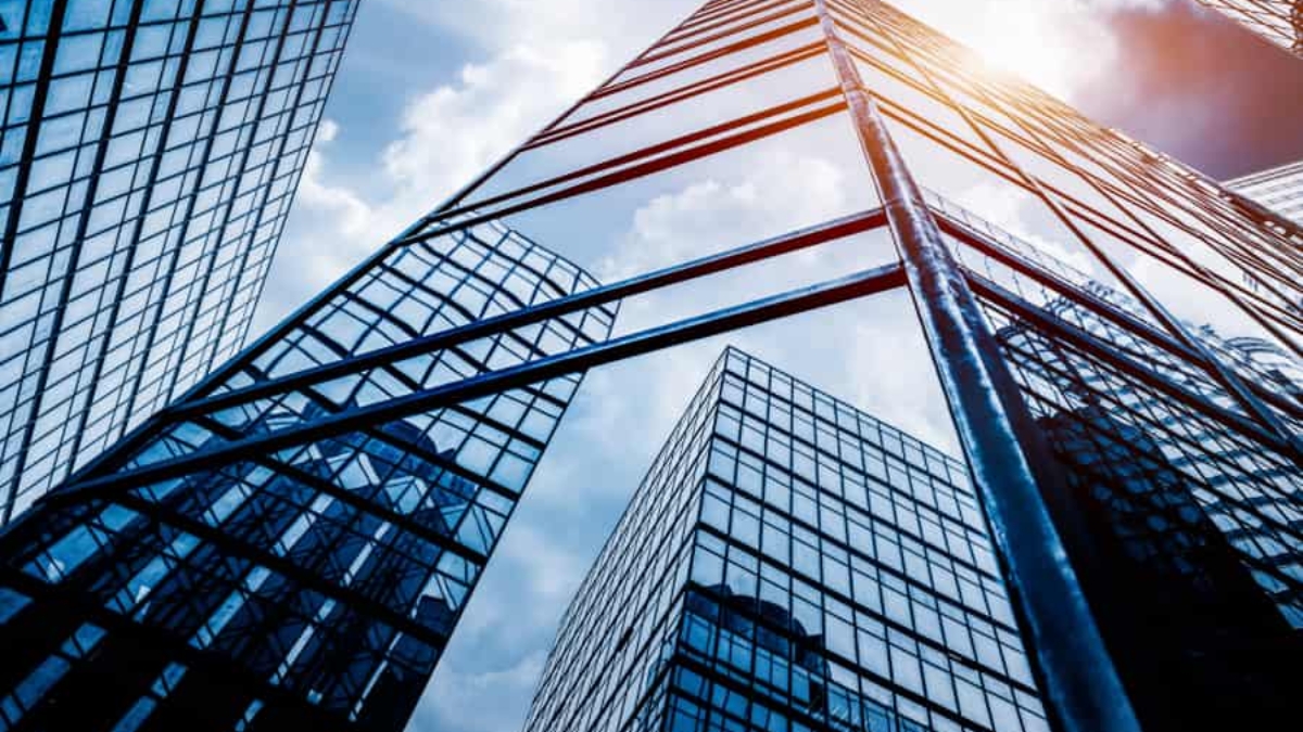 Tall glass skyscrapers reflect the blue sky and clouds as sunlight streams between the buildings, creating an urban cityscape with a modern architectural feel.