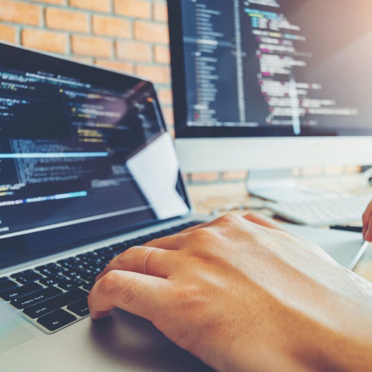 A person types on a laptop with programming code visible on the screen, possibly working on enVista Careers or a parcel audit service. Another monitor in the bright workspace, set against a brick wall, also displays code.