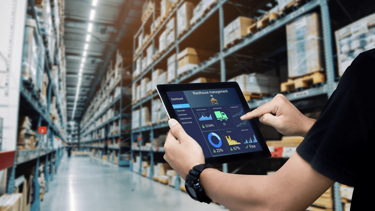 A person uses a tablet displaying warehouse management software in a large warehouse filled with shelves and boxes, highlighting digital inventory control and the benefits of warehouse automation technologies.