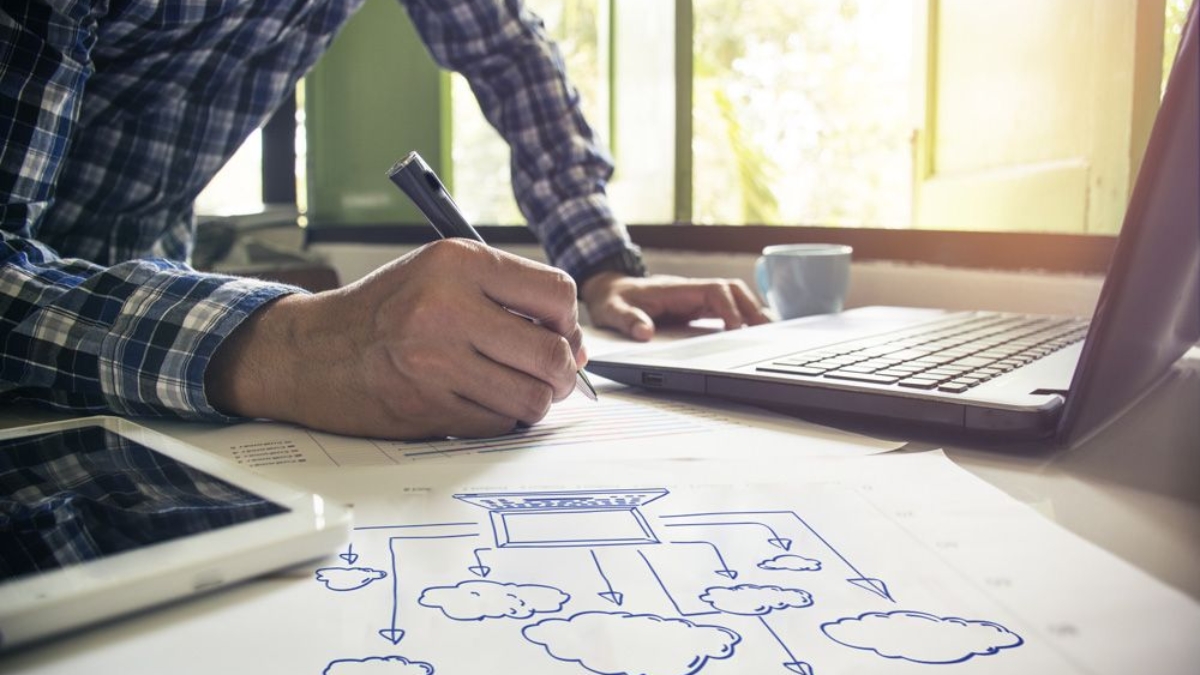 A person in a plaid shirt writes on paper next to a laptop, tablet, and a cloud computing diagram featuring D365 Business Central, with sunlight streaming through a window in the background.