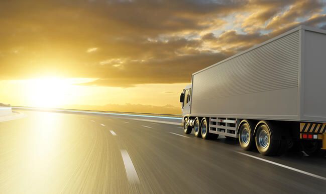 A large white semi-truck drives down an empty highway at sunset, with golden sunlight illuminating the road and clouds in the sky.