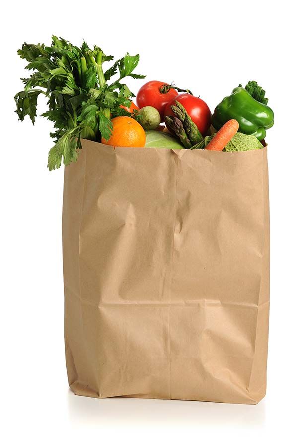 A brown paper grocery bag filled with fresh vegetables and fruits, including celery, tomatoes, green bell pepper, carrots, an orange, and asparagus—ready for transportation network optimization—against a white background.