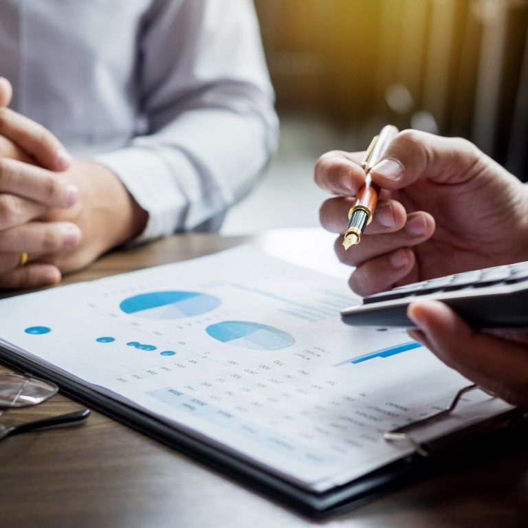 Two people sit at a desk reviewing financial charts on a clipboard, possibly discussing enVista 365 pricing. One holds a pen and calculator, while the other rests their hands together. Glasses and a small house model are also on the table.