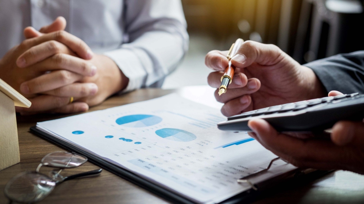 Two people sit at a desk reviewing financial charts on a clipboard, possibly discussing enVista 365 pricing. One holds a pen and calculator, while the other rests their hands together. Glasses and a small house model are also on the table.