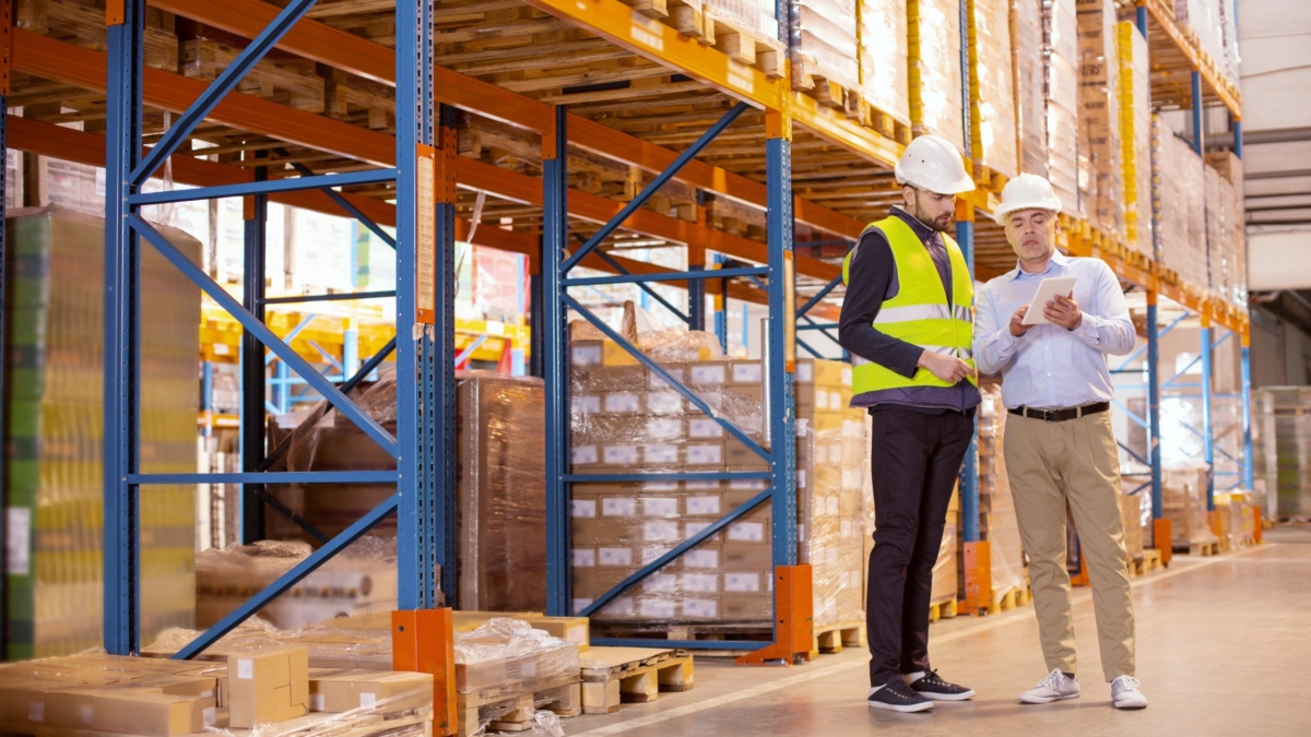 Two men in safety gear stand in a warehouse aisle lined with shelves and boxes. One wears a reflective vest and hard hat; the other holds a tablet, likely reviewing inventory using a labor management system as they discuss logistics.