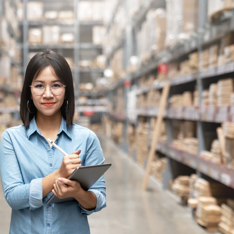 A woman with glasses and a blue shirt stands in a warehouse, holding a clipboard and pen, smiling at the camera. Shelves filled with boxes and supplies suggest efficient inventory management with Dynamics 365 Business Central.