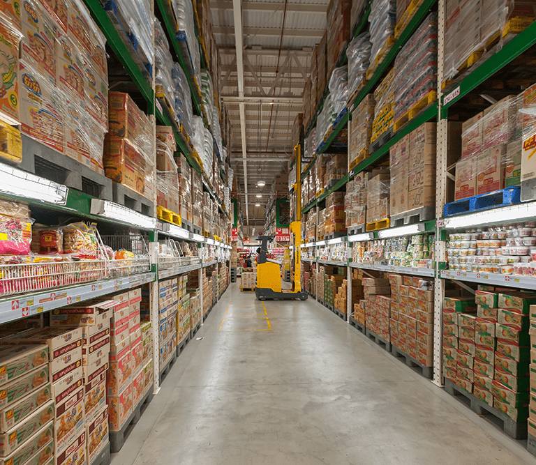 Warehouse aisle filled with stacked shelves of packaged goods, primarily instant noodles and food products; a worker operates a yellow pallet jack in the distance under bright overhead lighting.