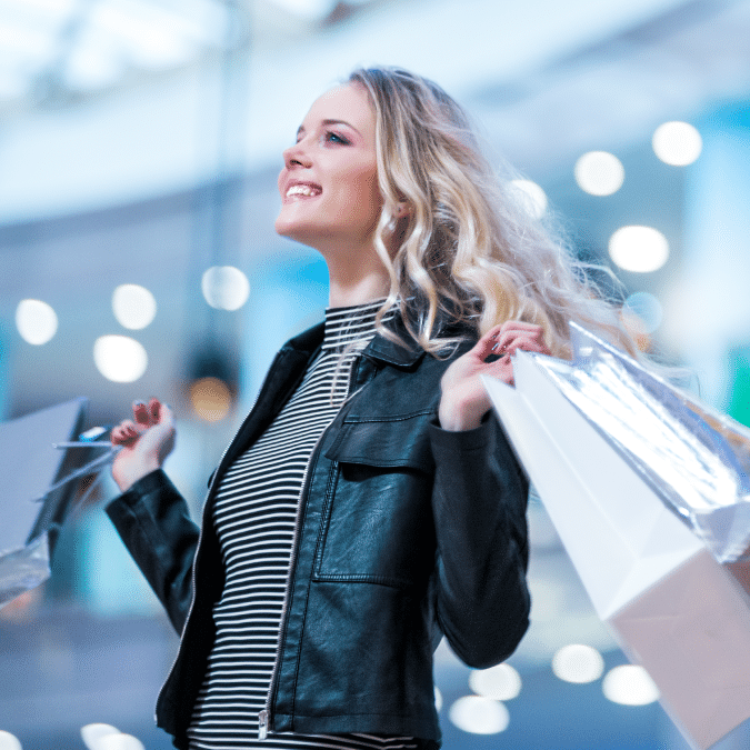 A smiling woman with long blonde hair, wearing a black leather jacket and striped shirt, holds several shopping bags in both hands. The background is blurred with bright lights, suggesting a mall or shopping center.