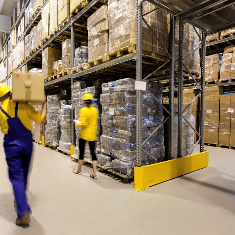 Two workers in safety helmets and uniforms move boxes in a large warehouse with tall shelves stacked with boxes and pallets. The warehouse is brightly lit and organized, with wide aisles for easy movement.
