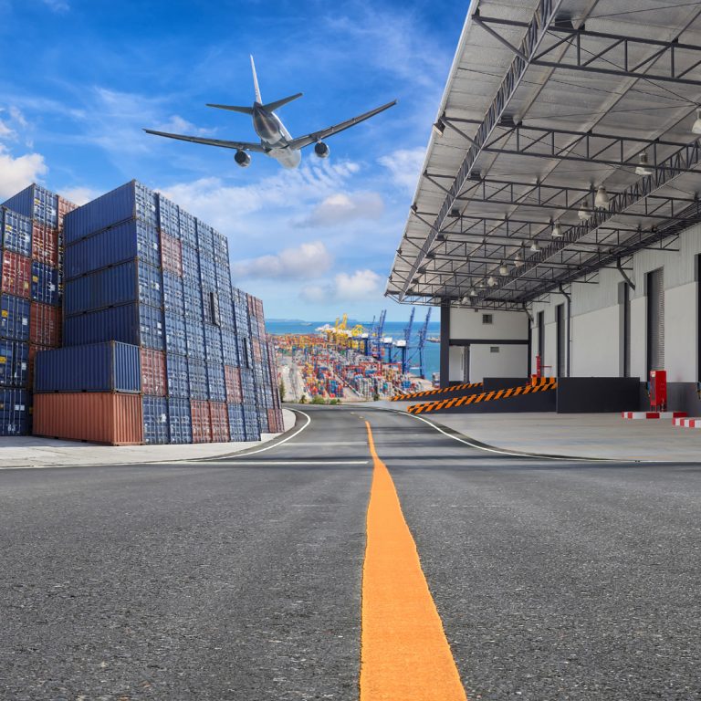 A cargo plane flies over stacked shipping containers near a warehouse at an industrial port, with a clear sky and road marking leading toward the buildings.
