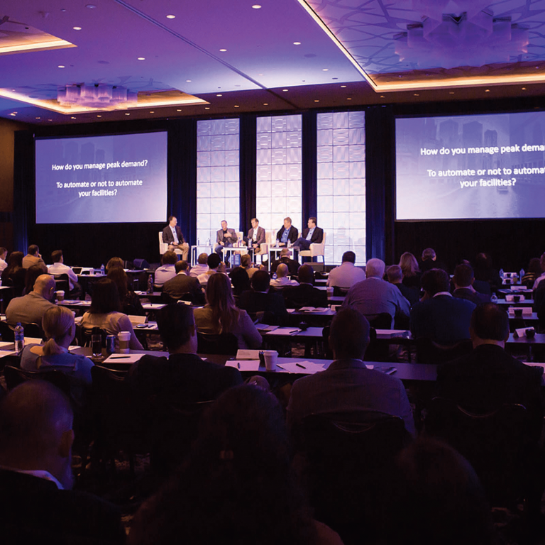 A large audience watches a panel discussion in a conference room. Four speakers sit onstage in front of three large screens displaying the question, “How do you manage peak demand? To automate or not to automate your facilities?”.