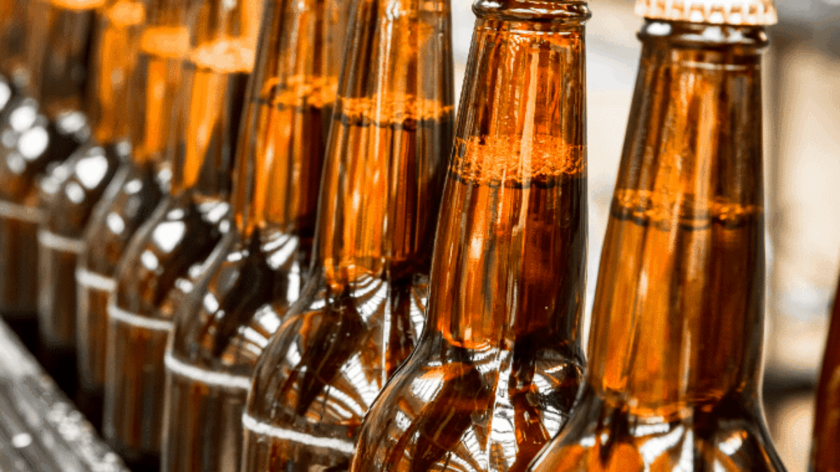 Close-up of amber glass beer bottles with caps, lined up in a row on a conveyor belt in a brewery or bottling facility.
