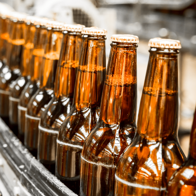 Close-up of amber glass beer bottles with caps, lined up in a row on a conveyor belt in a brewery or bottling facility.