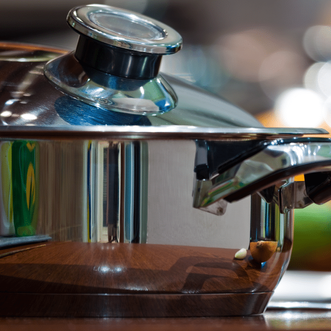 A close-up of a shiny stainless steel pot with a glass lid, sitting on a wooden surface. The pots handle and lid knob are visible, reflecting colorful shapes from the surrounding kitchen.