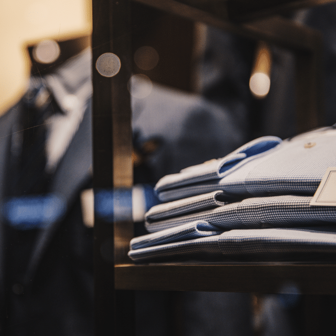 A close-up of neatly folded blue dress shirts displayed on a shelf in a clothing store, with blurred suits and garments in the background.