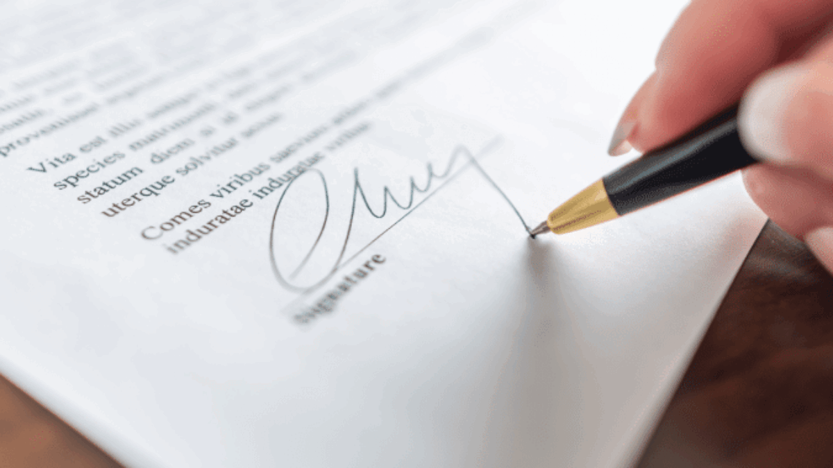 A close-up of a person signing a document with a black and gold pen. The signature is being written at the bottom of the page, and the text above is slightly blurred.