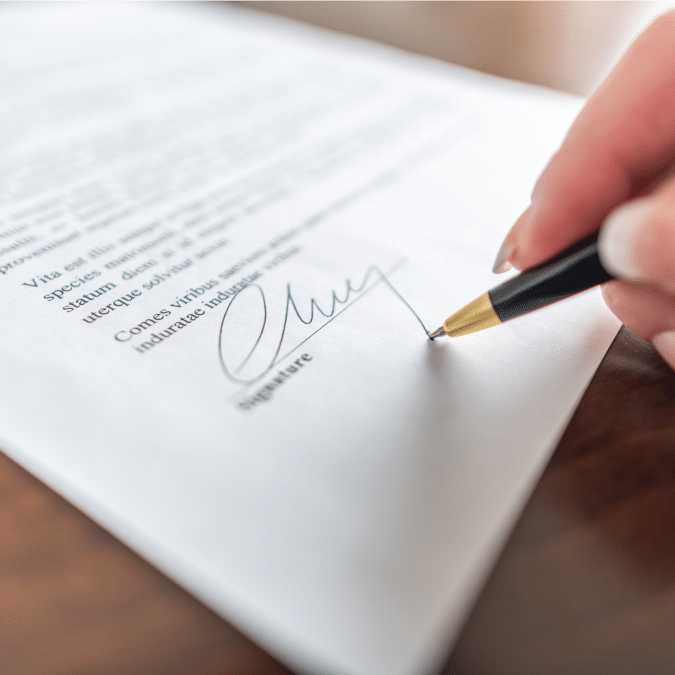 A close-up of a person signing a document with a black and gold pen. The signature is being written at the bottom of the page, and the text above is slightly blurred.