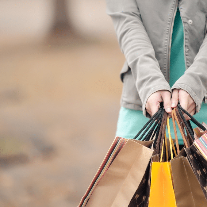 A person in a grey jacket and teal dress holds several colorful shopping bags in both hands, standing outdoors on a blurred background.