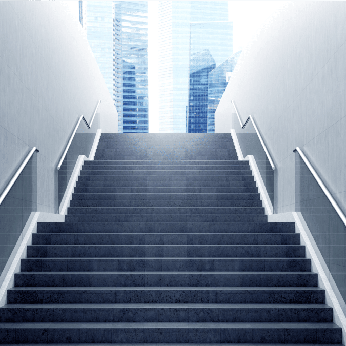 A wide, modern staircase with metal railings on both sides leads upward to bright daylight, with tall glass office buildings visible in the background.
