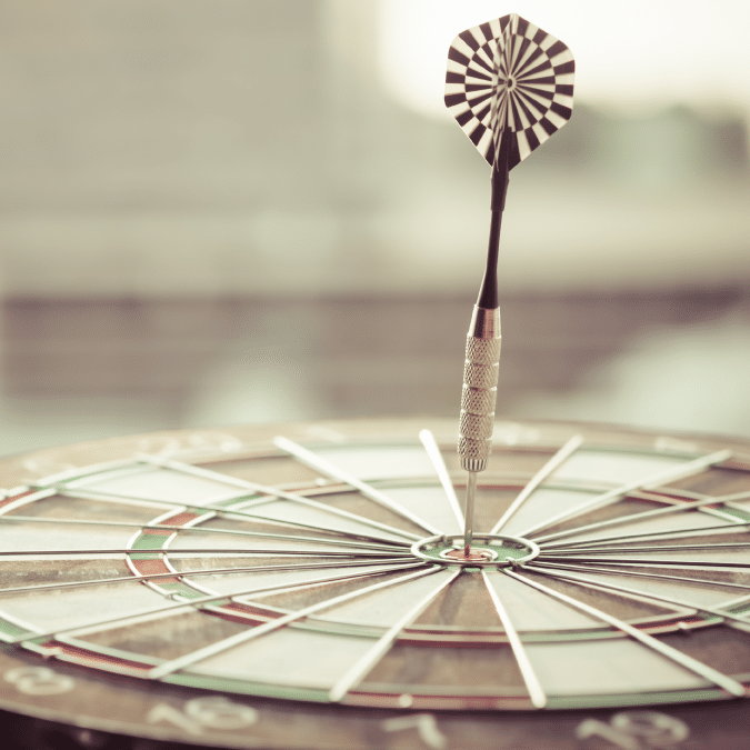 A dart is precisely embedded in the bullseye of a dartboard, illustrating accuracy and focus; the background is softly blurred, keeping the attention on the dart and the target.