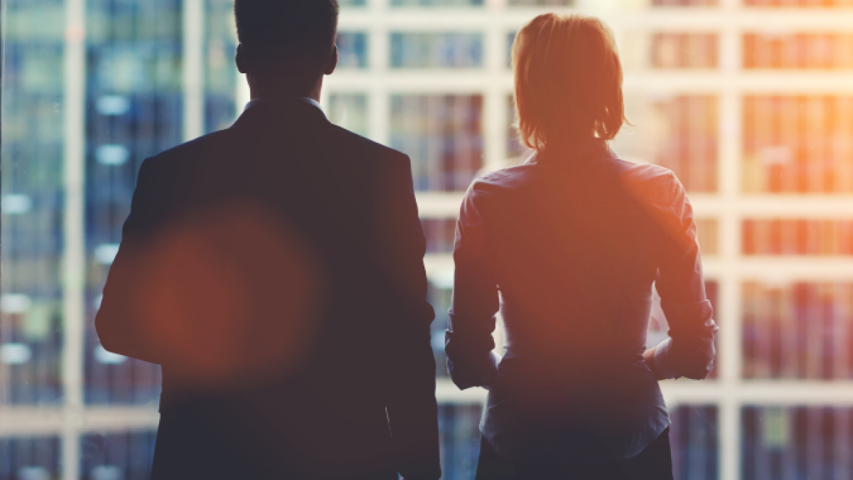 Two people in business attire stand side by side, silhouetted against a large office window with a cityscape of tall buildings in the background, sunlight streaming in.
