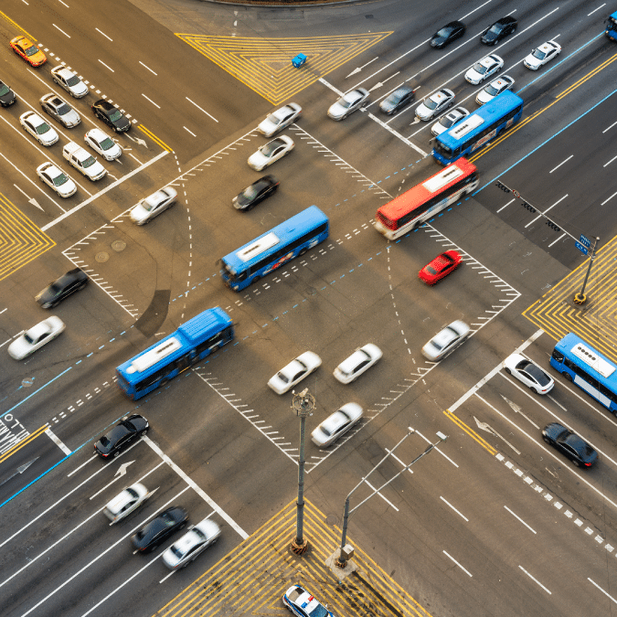 Aerial view of a busy city intersection with cars, buses, and a red vehicle moving in multiple directions, traffic lanes and markings clearly visible.