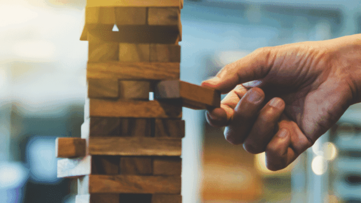 A hand carefully pulls a wooden block from a stacked Jenga tower, with blurred indoor background and sunlight streaming in from the left.