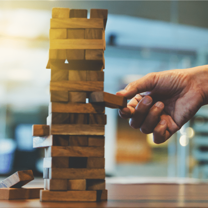 A hand carefully pulls a wooden block from a stacked Jenga tower, with blurred indoor background and sunlight streaming in from the left.
