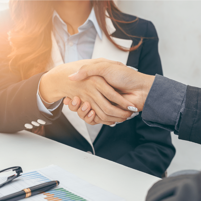 Two people in business attire shaking hands over a desk with documents, a pen, and eyeglasses, suggesting a successful meeting or agreement.