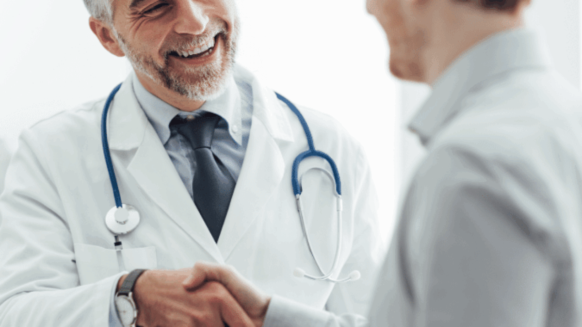 A smiling male doctor with gray hair and a beard, wearing a white coat and stethoscope, shakes hands with a male patient in an office or clinical setting.
