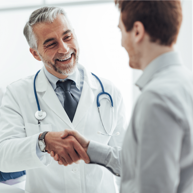 A smiling male doctor with gray hair and a beard, wearing a white coat and stethoscope, shakes hands with a male patient in an office or clinical setting.