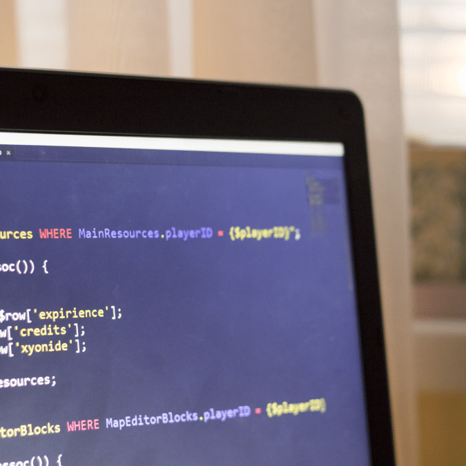A close-up of a computer screen displaying code, with part of a laptop bezel visible. The code appears to be written in SQL and another programming language. Blurred curtains and sunlight are in the background.