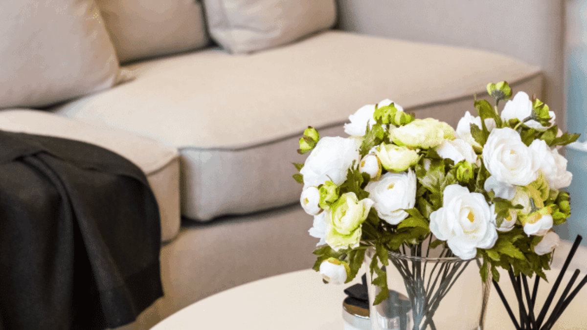 A beige sofa with a dark blanket draped over the armrest sits behind a round coffee table, which holds a glass vase of white and yellow flowers, a candle, and a reed diffuser.