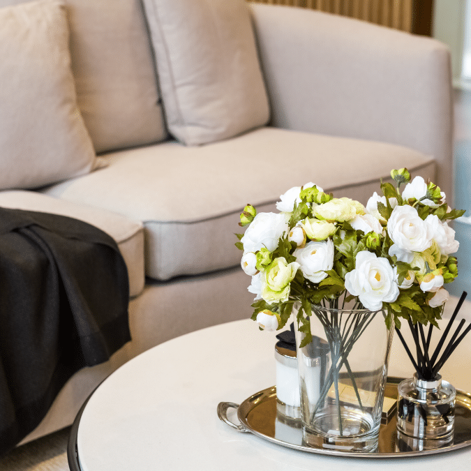 A beige sofa with a dark blanket draped over the armrest sits behind a round coffee table, which holds a glass vase of white and yellow flowers, a candle, and a reed diffuser.