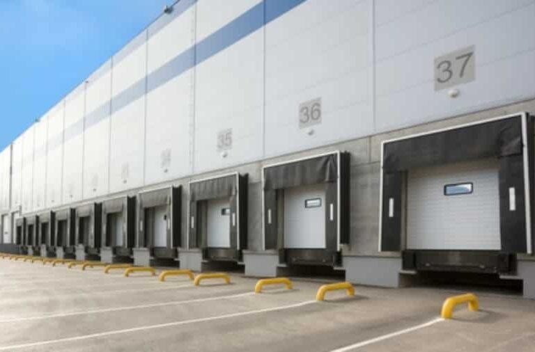 A row of loading docks with numbered bays and closed overhead doors at a large, modern warehouse under a clear blue sky.