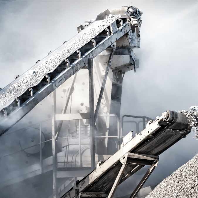 Industrial conveyor belts transport gravel amid clouds of dust at a quarry or mining site, with metal structures and machinery visible in the background.