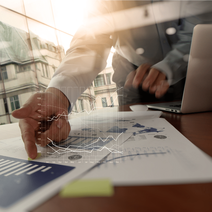 A person points at financial documents and graphs on a desk, with a laptop nearby and a virtual graph overlay, suggesting data analysis or business strategy in a modern office setting.