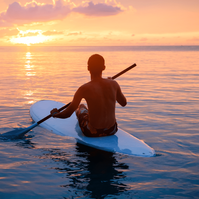 A person paddles on a stand-up paddleboard in calm ocean waters at sunset, with warm orange and pink hues in the sky and sunlight reflecting on the water’s surface.