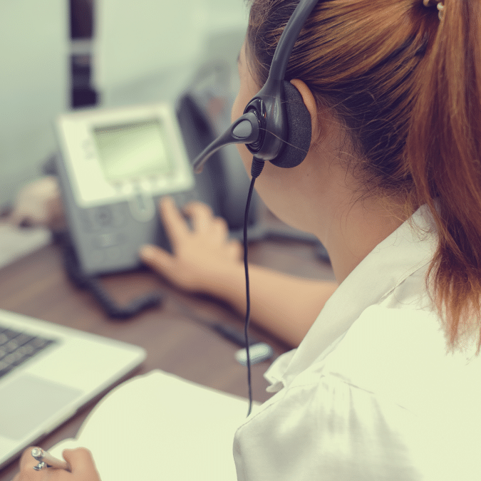 A woman wearing a headset sits at a desk, using a telephone while looking at a computer screen and holding a pen, suggesting she is working in a customer service or call center environment.