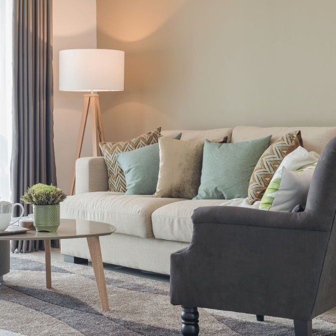 A cozy living room with a beige sofa decorated with green and patterned pillows, a dark gray armchair, a wooden coffee table with a small plant and mugs, a standing lamp, and floor-length curtains.