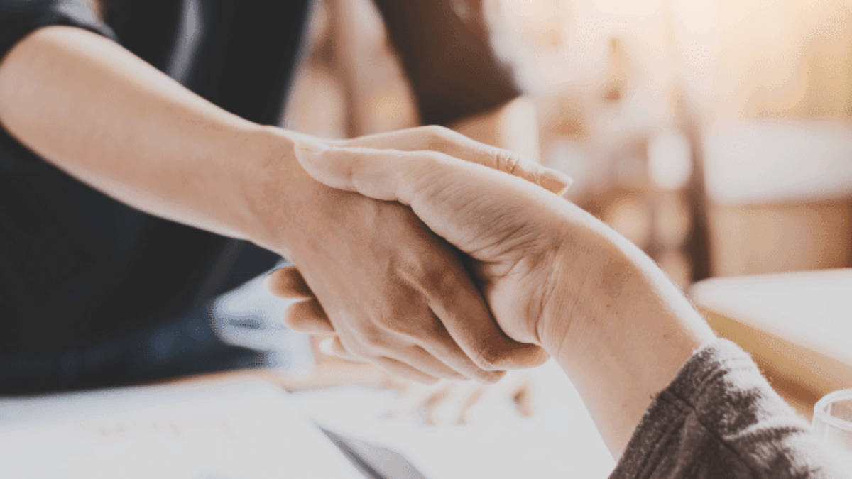 Two people shaking hands across a desk with documents and a pen, suggesting a business agreement or partnership in a professional setting.