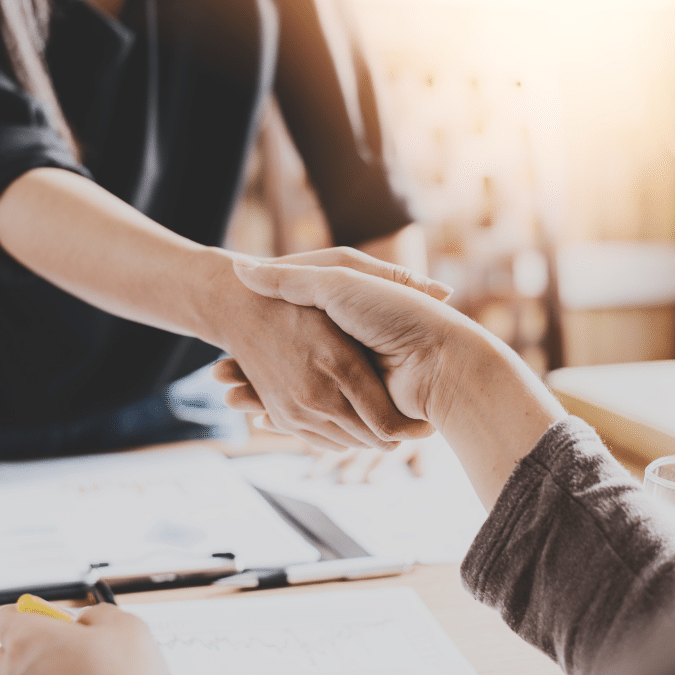 Two people shaking hands across a desk with documents and a pen, suggesting a business agreement or partnership in a professional setting.