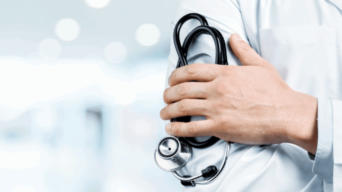 A doctor wearing a white lab coat stands with arms crossed, holding a stethoscope in one hand. The background is blurred, suggesting a hospital or medical facility setting.
