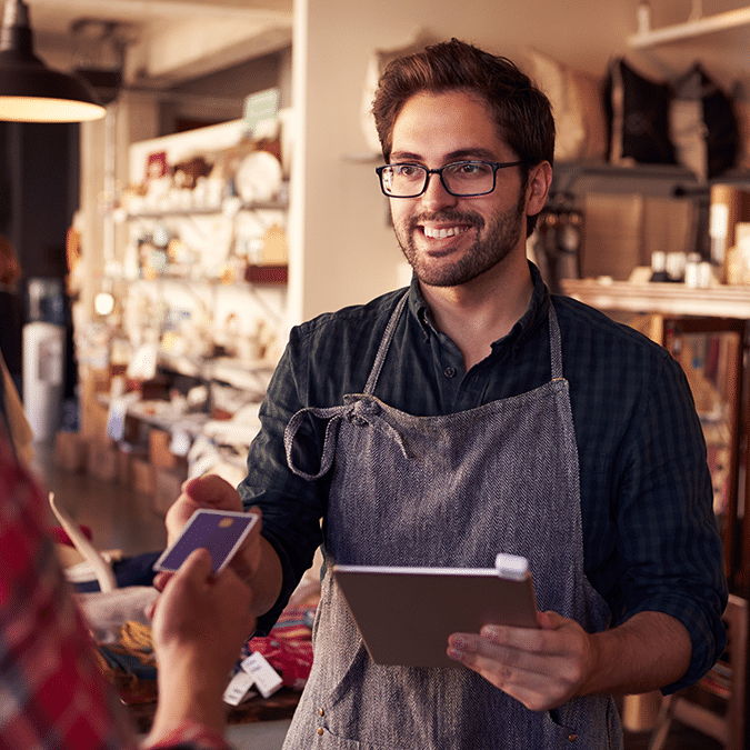 A smiling shop employee wearing an apron and glasses holds a tablet while a customer hands him a credit card in a cozy, well-lit store.