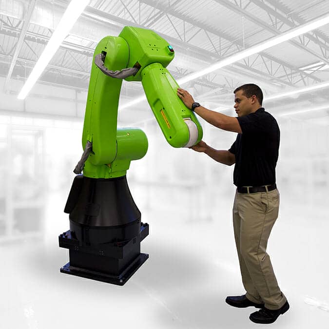 A man in khaki pants and a black shirt interacts with a large green industrial robotic arm in a bright, modern factory setting.