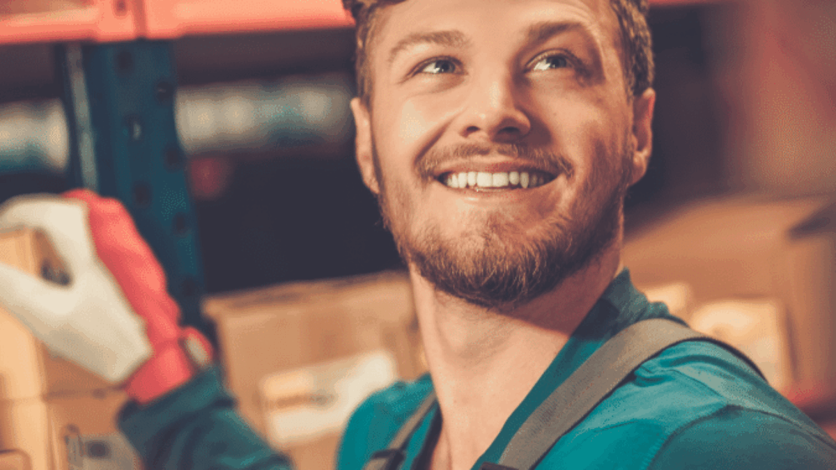 A young man wearing work gloves and an apron smiles while organizing boxes on shelves in a warehouse or storage area.