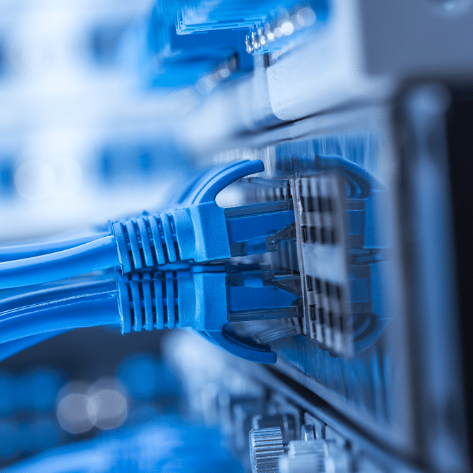 Close-up of several blue Ethernet cables plugged into a network switch, with blurred electronic components in the background, depicting a data center or server room environment.