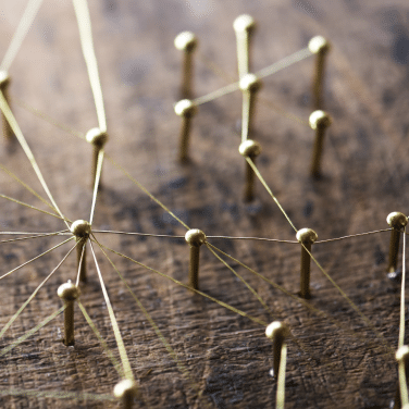 Close-up of golden pins connected by thin threads, arranged on a wooden surface to form a network or web-like pattern representing interconnectedness.