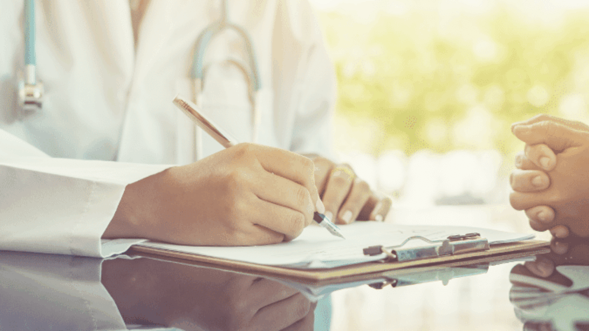 A doctor in a white coat writes on a clipboard while holding hands with a patient, offering comfort and support during a consultation at a medical office.